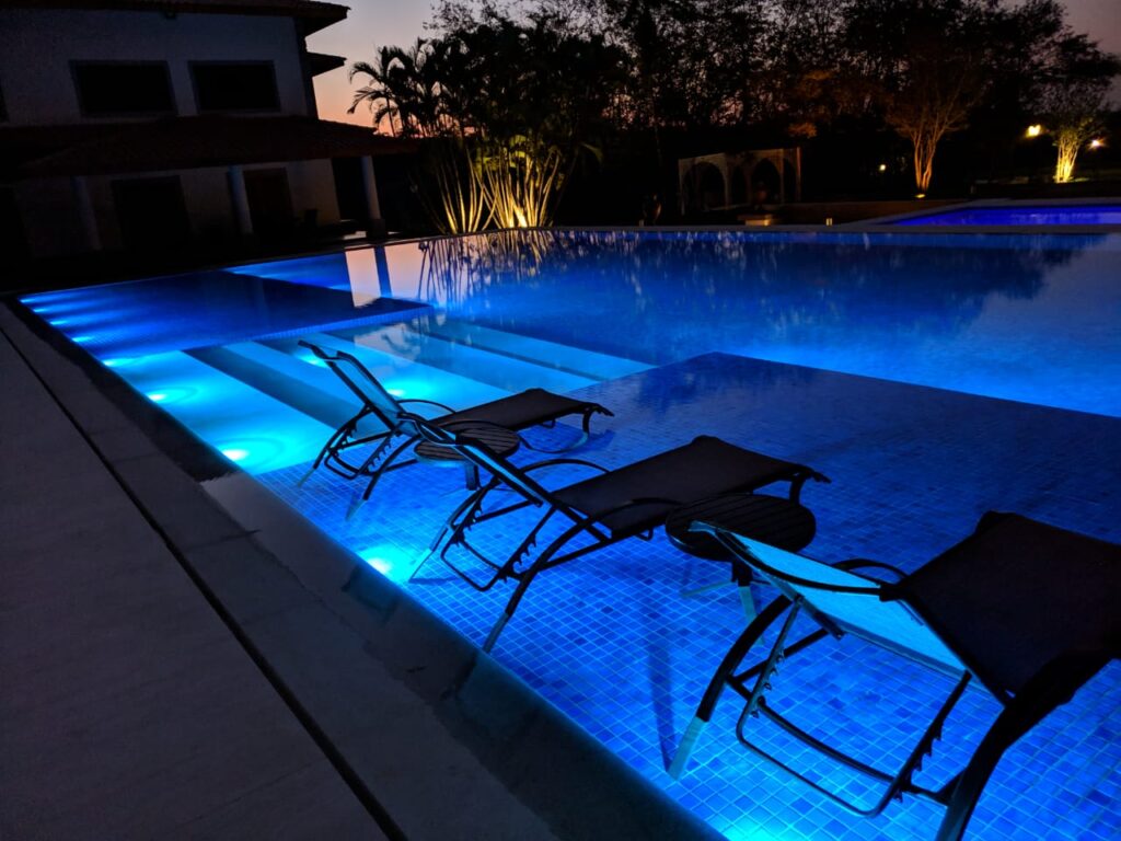 Piscina de luxo iluminada com refletores LED subaquáticos na cor azul, destacando espreguiçadeiras parcialmente submersas e o design arquitetônico refinado do ambiente. A imagem evidencia o impacto visual e a sofisticação que a iluminação premium da Cristal Led & Inox proporciona em projetos residenciais de alto padrão.